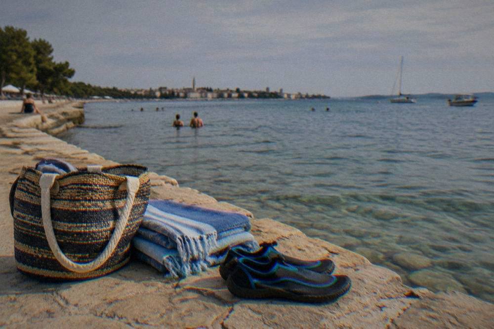 A beach bag, folded towel and water shoes on a stone ledge beside a clear shallow sea.