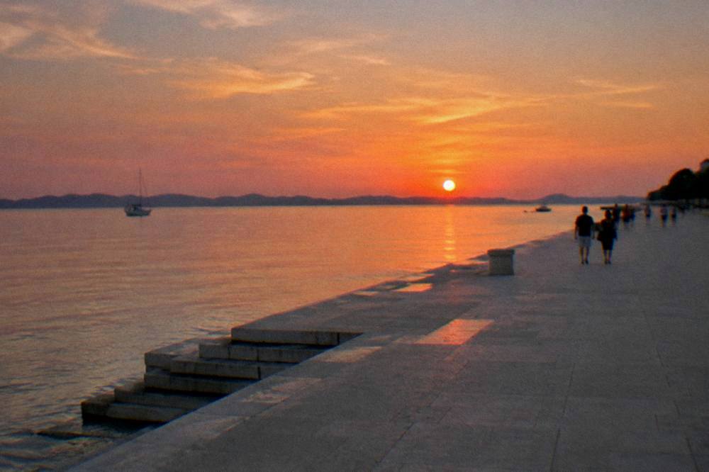 The sunset over the Adriatic from a seaside promenade with a few distant walkers.