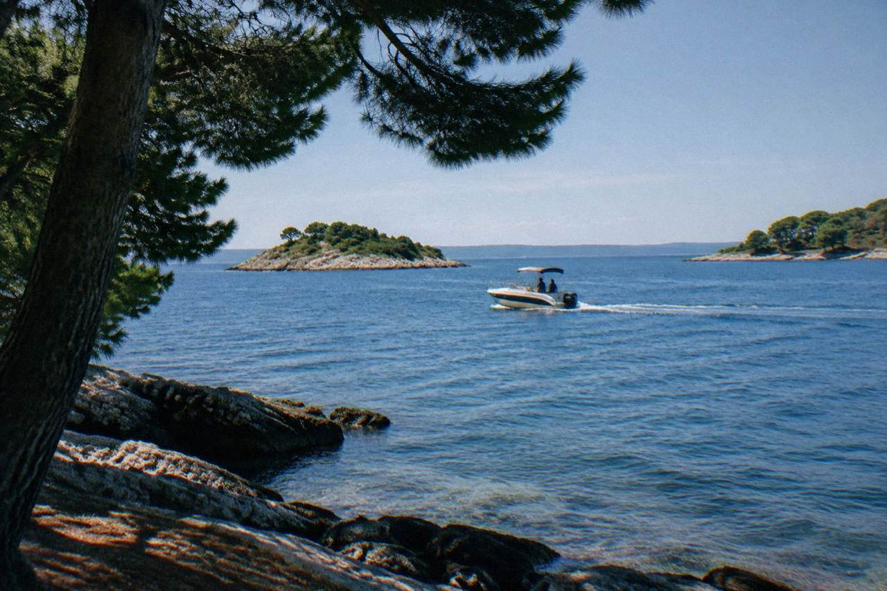 A small boat cruising on calm blue water near rocky islets backed by pines.