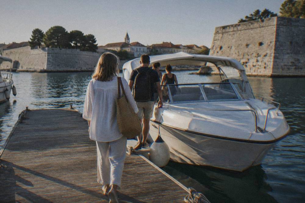 Passengers seen from behind boarding a white speedboat at a wooden dock in a calm harbour.