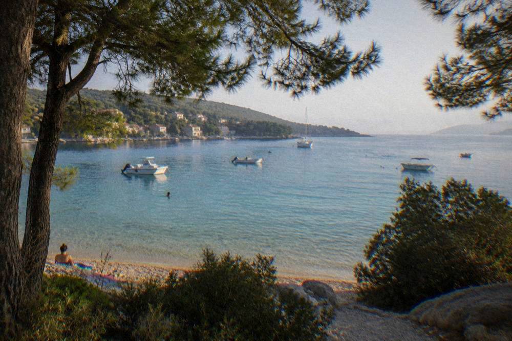 Turquoise shallows at a sandy lagoon beach framed by pines with small boats anchored.