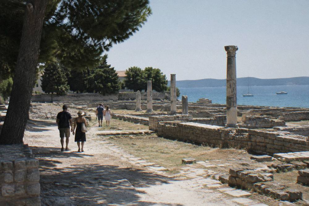 Distant visitors walking past weathered stone ruins of the Roman Forum in bright sun.