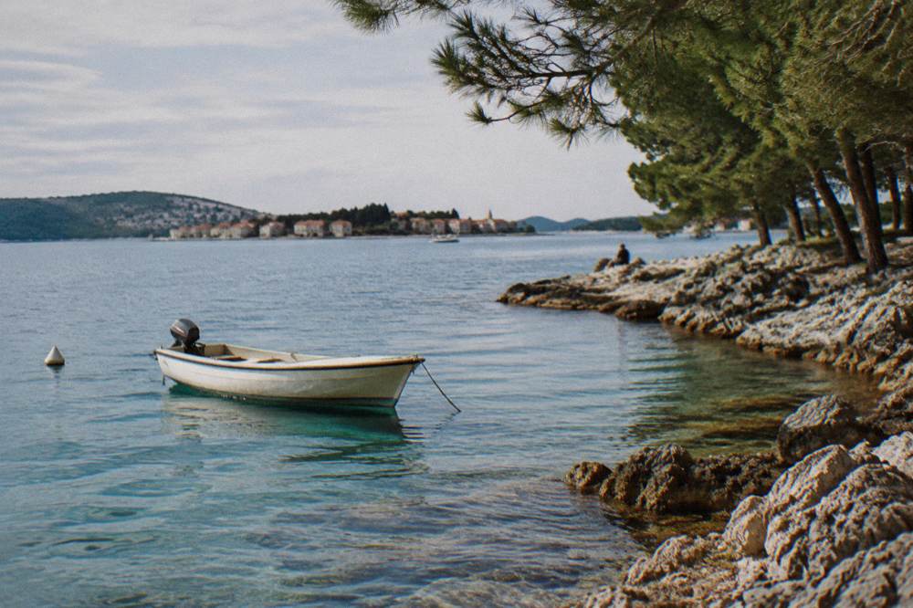 A small boat anchored in clear turquoise water beside a pine-covered rocky shore.