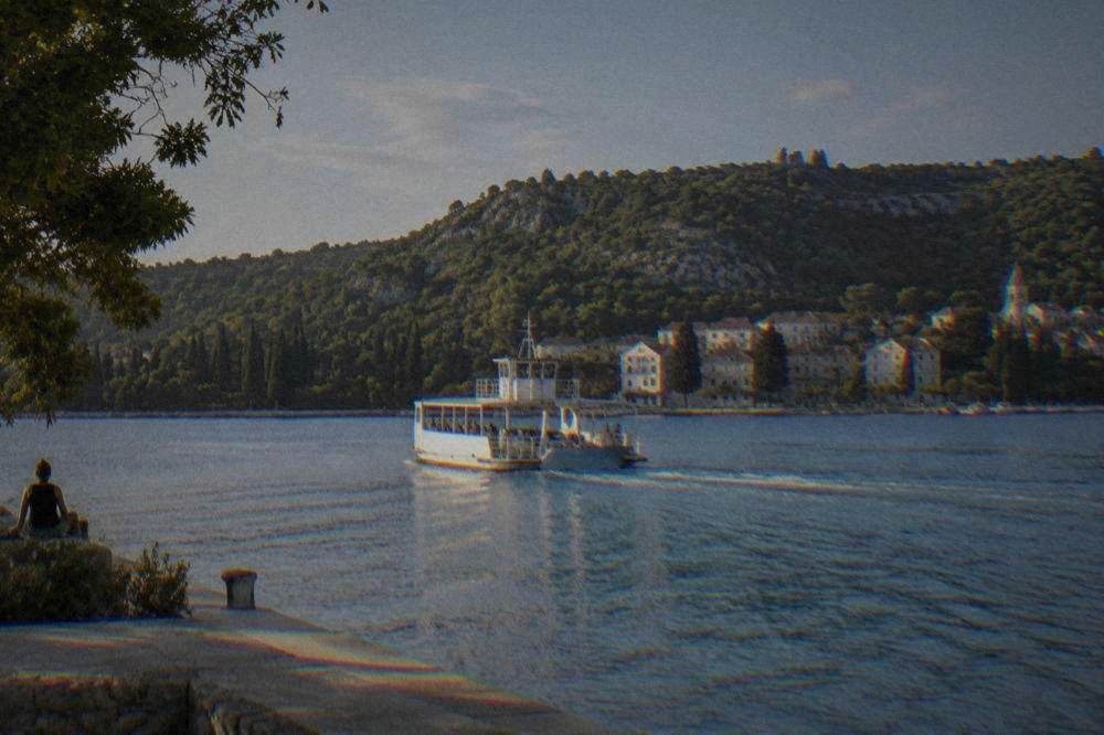 A small white ferry crossing calm blue water toward a wooded shore and a river town.