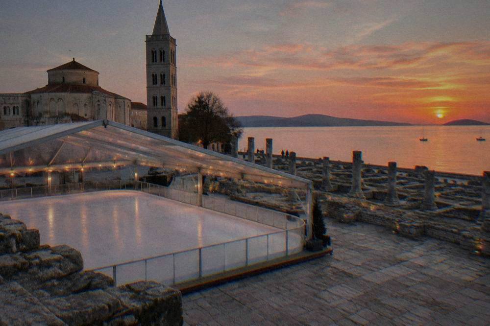 A temporary covered ice rink beside Roman ruins on an old stone square at dusk.