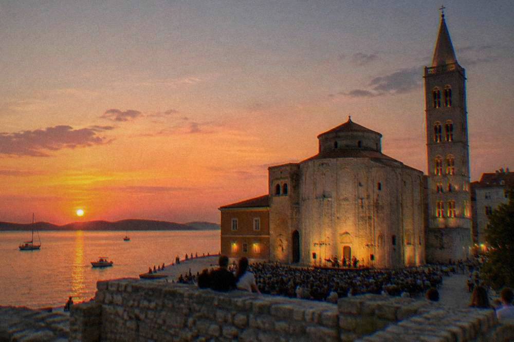 A distant evening crowd in front of a lit stone church facade with a bell tower.