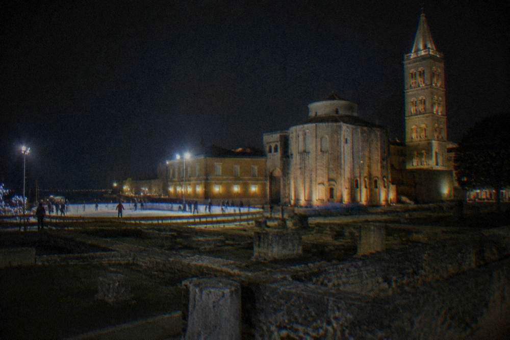 Distant skaters on a temporary rink beside floodlit Roman ruins at night.