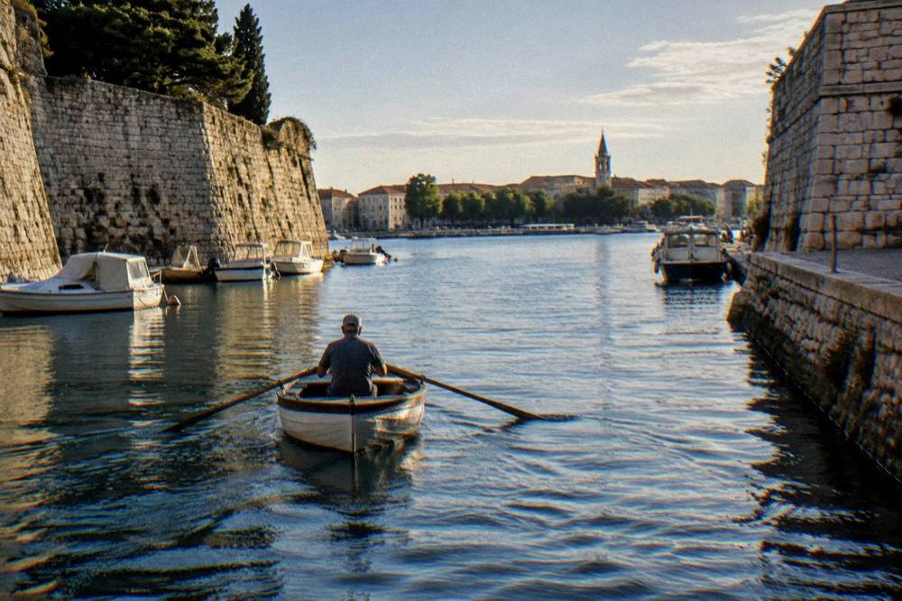A small wooden rowing boat crossing a narrow harbour channel between stone walls.