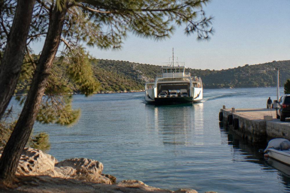 A car ferry approaching an island jetty between pine trees on calm water.