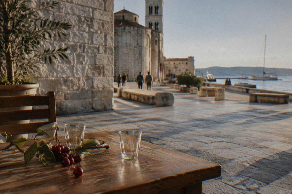 Two small empty glasses and cherries on a wooden cafe table at a stone-walled corner in Zadar.