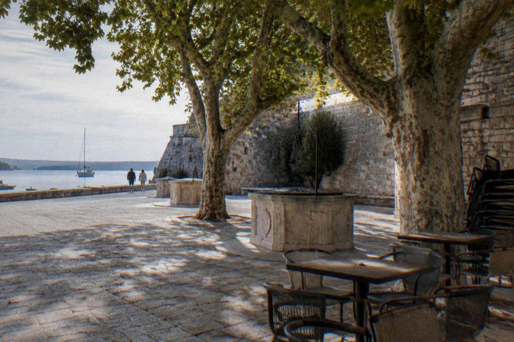 Calm cafe corner at Five Wells Square in Zadar with plane trees and stone wall.