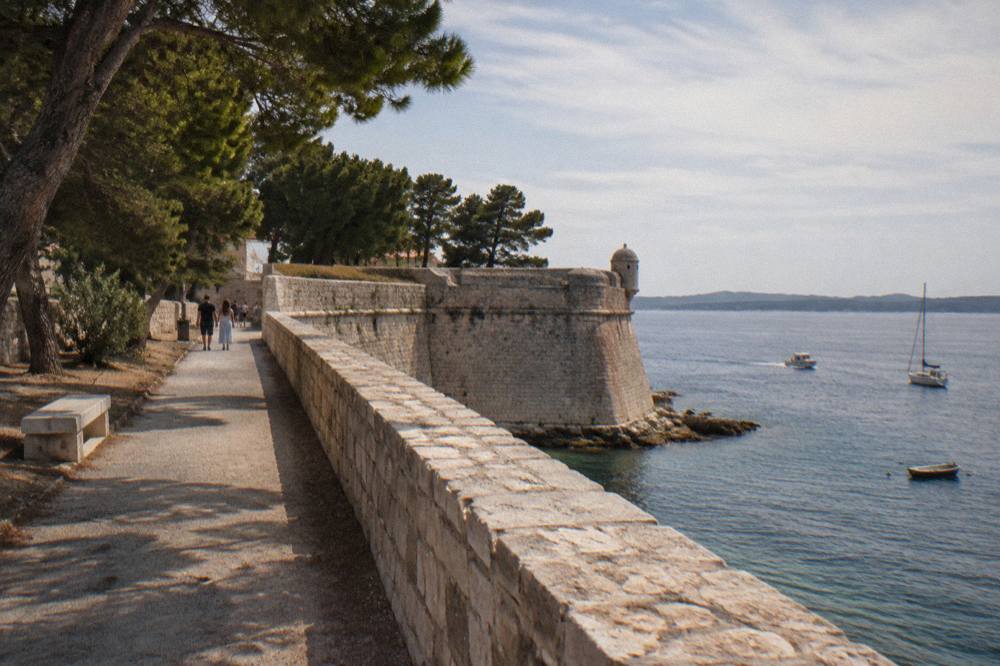 Weathered limestone city walls and a bastion with pine trees beside the path in Zadar.