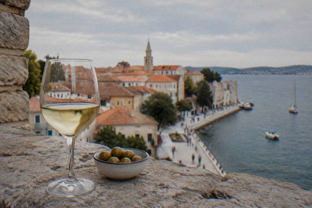 A wine glass and olives on a stone ledge with terracotta roofs behind in Zadar.