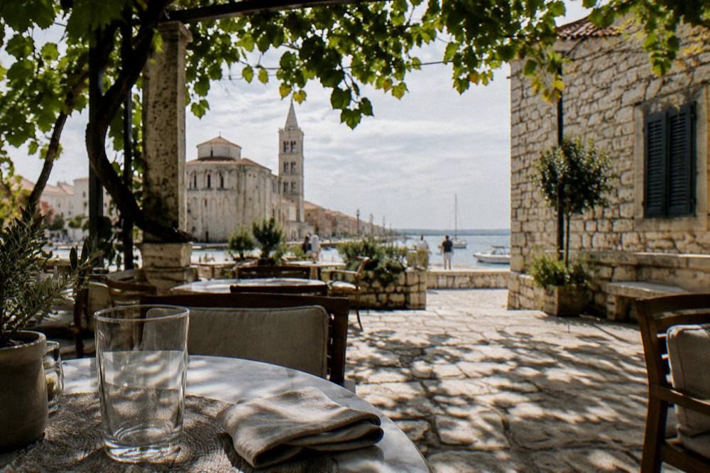 A cafe table under a vine pergola in a Zadar old-town courtyard in early autumn light.