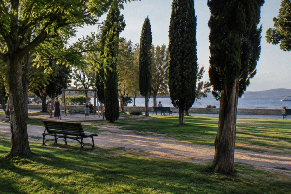 A shaded park lawn with benches and cypress trees in Zadar on a calm day.