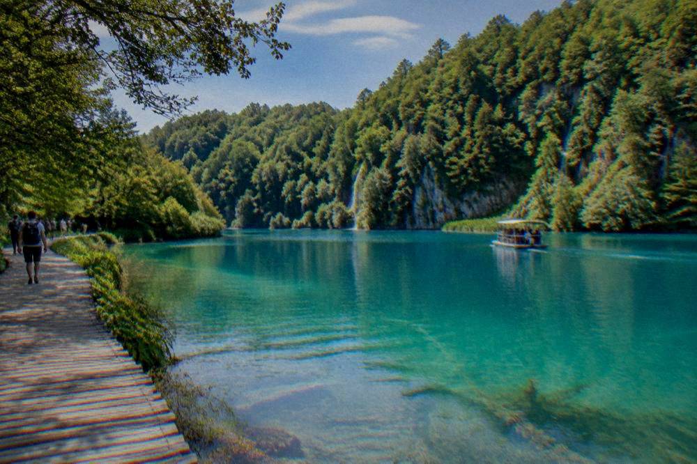 A turquoise lake at Plitvice with a wooden walkway beside tall forest.