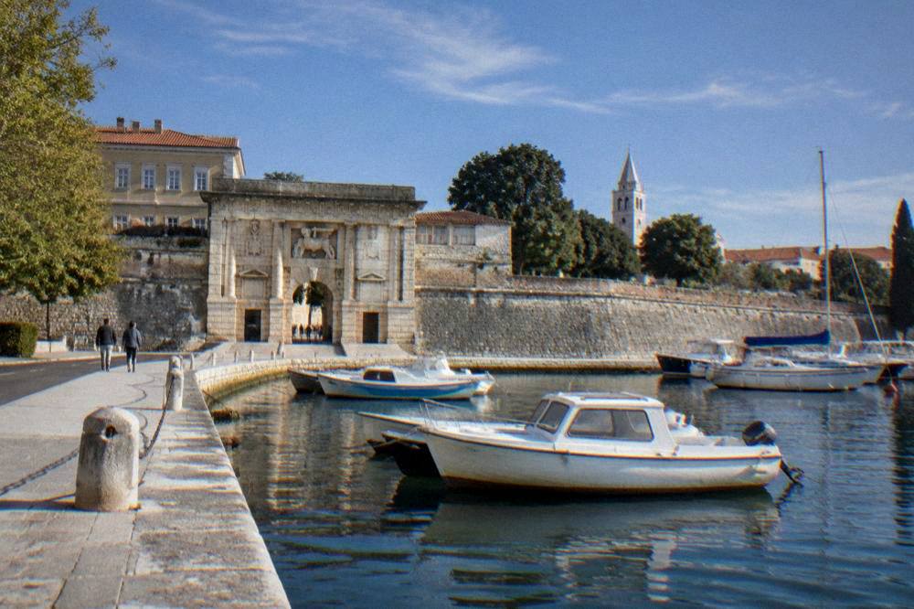 Land Gate beside the small harbour with moored boats in Zadar, walkers far away.