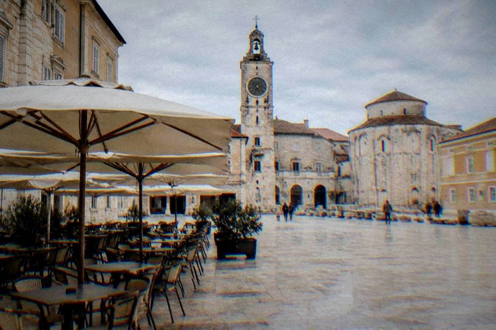 Stone square cafe with empty chairs under parasols and a clock tower