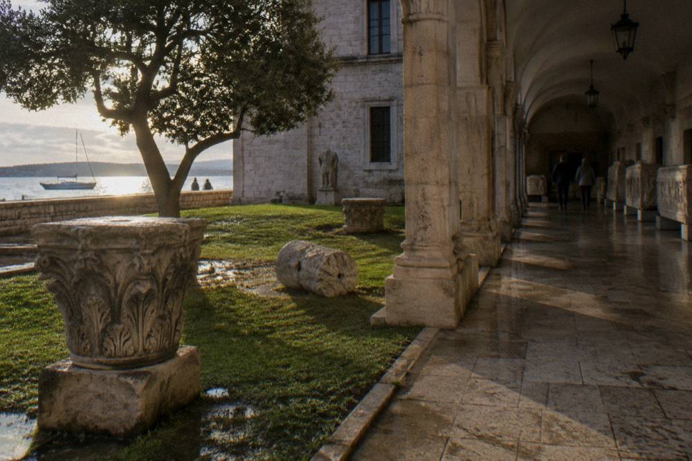 Stone courtyard arcade with carved fragments on damp grass on a clear day