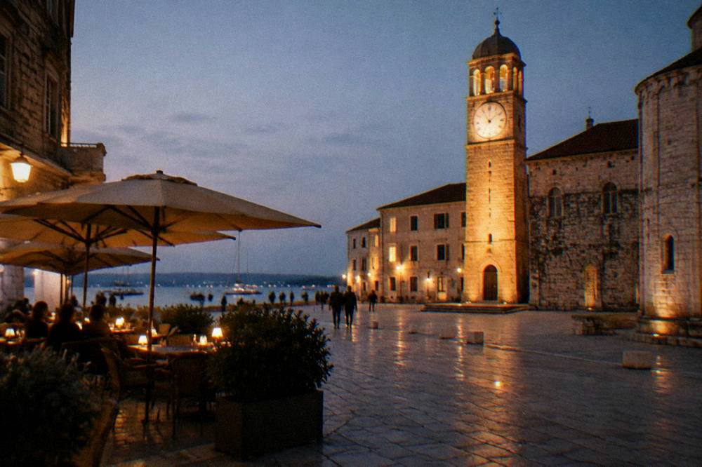 Square cafe terrace at night with lit tables and a glowing clock tower